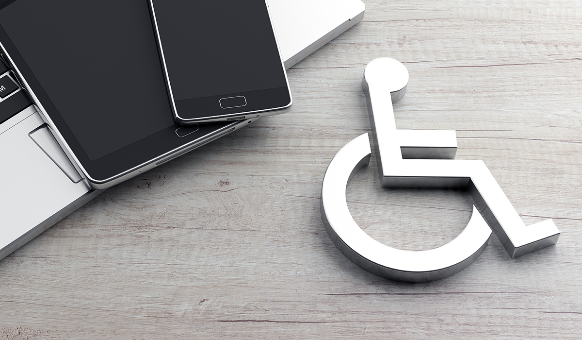 This image features a composition that includes a laptop, a smartphone, and a tablet, all lying on a flat wooden surface with their screens facing up. They are placed in a slightly fanned-out arrangement on the left side of the frame. To the right of the devices, there's a large metallic icon of the International Symbol of Access, commonly known as the wheelchair symbol, signifying accessibility. It's depicted in 3D, casting a slight shadow on the wooden surface, indicating that the symbol is standing upright. The overall image seems to convey a message about the importance of accessibility in technology.