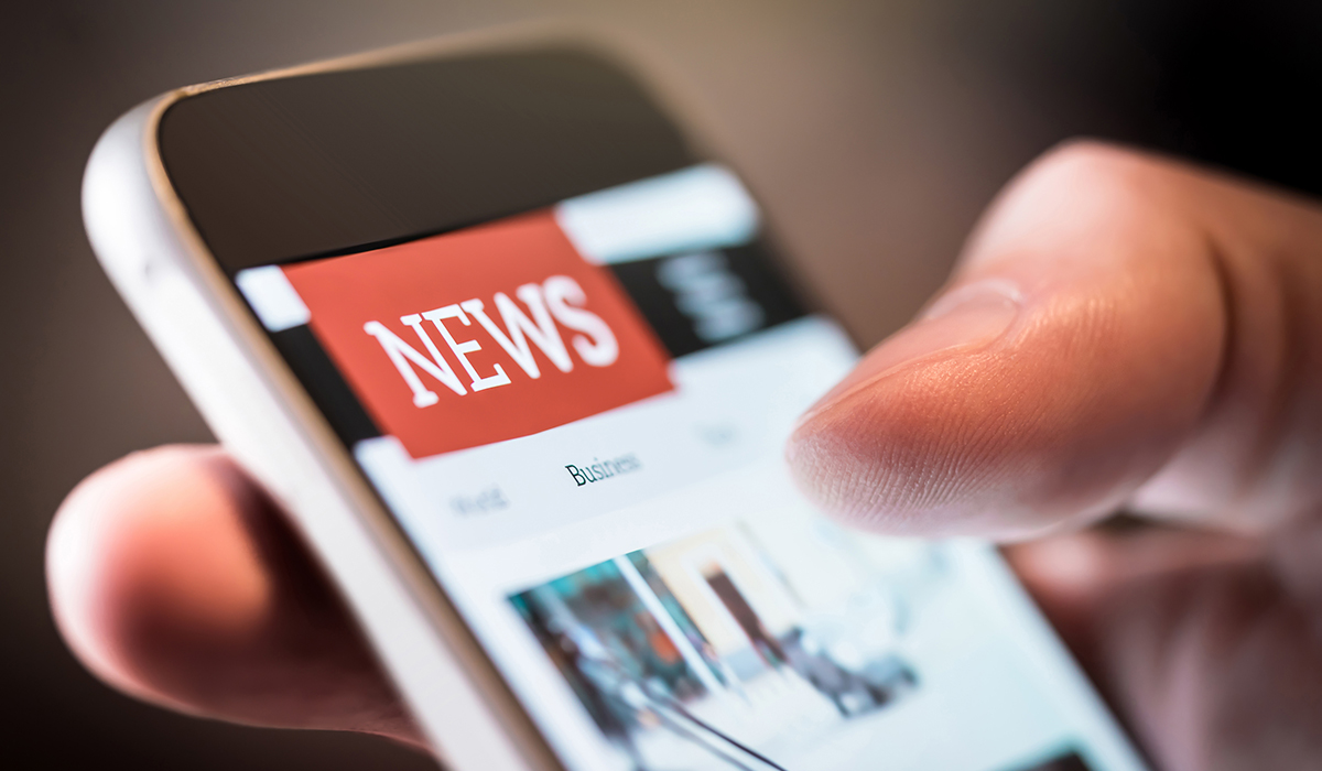 The image is a close-up photo of a person's hand holding a smartphone. The screen of the phone is in clear focus and displays a news application interface. Prominently at the top of the screen is a bold red rectangle with the word "NEWS" in white capital letters. Below this banner are tab options, such as "World", "Business", and other categories not fully visible. The person's thumb is extended and about to interact with the touch screen, perhaps to scroll through the news articles. The lighting on the thumb indicates a warm environment, possibly indoors. The phone and the hand occupy the center and foreground of the image, and the background is out of focus, emphasizing the action of reading the news on the phone.