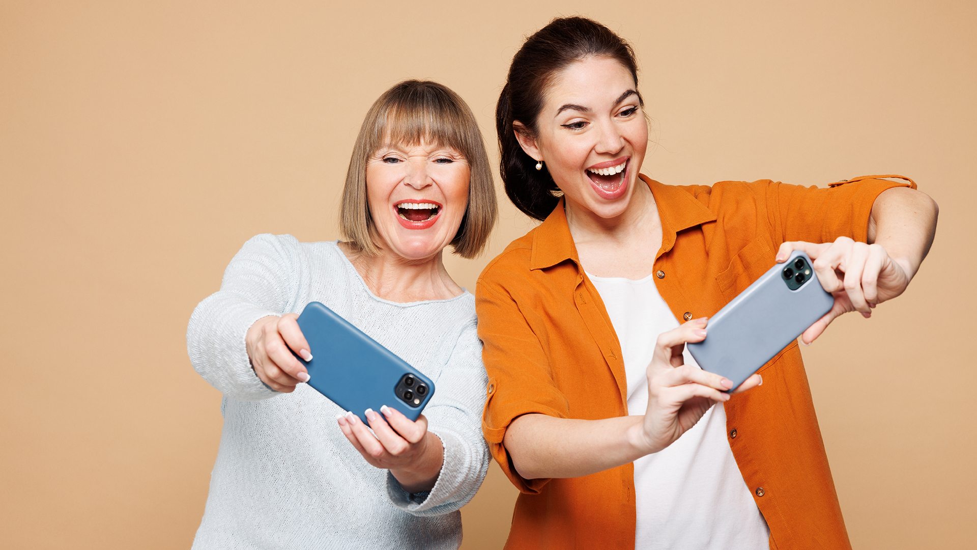 Two women are joyfully taking selfies against a plain beige background, both smiling widely with excited expressions; the older woman on the left has straight, shoulder-length blonde hair and wears a light gray sweater while holding a blue phone, and the younger woman on the right has dark hair pulled back, wears an orange shirt over a white top, and holds a gray phone.